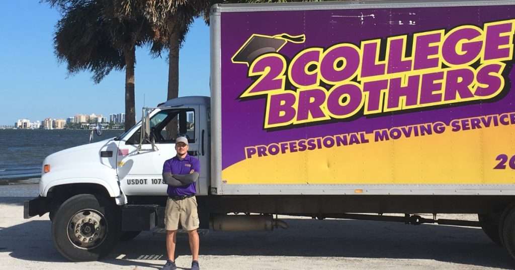 A professional 2 College Brothers mover standing proudly with his arms crossed in front of a branded purple and yellow moving truck parked near a sunny waterfront with palm trees.